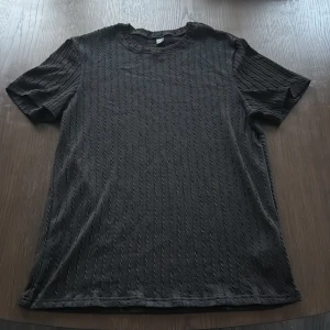 Svart ribbad t-shirt från Manfinity - Svart t-shirt från Manfinity i storlek L med ribbad struktur och diskret kabelstickat mönster. T-shirten har rund halsringning och korta ärmar, perfekt för en clean och stilren look. Materialet känns mjukt och följsamt mot huden.
