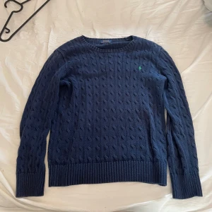 Mörkblå kabelstickad tröja Polo Ralph Lauren - Kabelstickad mörkblå tröja från Polo Ralph Lauren med rund halsringning och grönt broderat logotyp på bröstet. Tröjan har långa ärmar och breda ribbade muddar vid ärmslut och nederkant. Perfekt för lager-på-lager-stil. Storlek XL men sitter som M. Nypris 2399kr