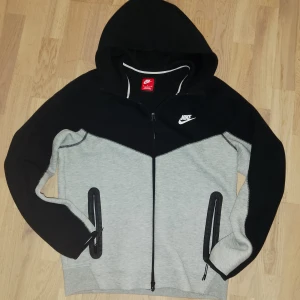 Nike Tech hoodiei svart och grå, M - Nike hoodie i svart och ljusgrått med hel dragkedja och huva. Tröjan har två fickor med dragkedja och klassisk Nike-logga på bröstet. Perfekt för chill eller träning, riktigt skön och sportig stil.