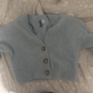 Ljusblå kofta från DIVIDED XS - Superfin ljusblå kofta från DIVIDED i stickad material. Croppad modell med ribbad struktur och tre snygga träknappar framtill. Perfekt för att ge din outfit en chill vibe. Passar dig som gillar mjuka och mysiga plagg.