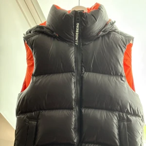 Svart dunväst från J.Lindeberg - Snygg svart dunväst från J.Lindeberg med orange insida och hög krage. Västen har en tydlig logotyp på dragkedjan och insidan, samt praktiska fickor. Perfekt för lager-på-lager och har en modern, puffad look.