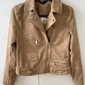 Beige mockajacka från H&M Divided - Snygg beige bikerinspirerad jacka i mockaimitation från H&M Divided. Jackan har guldiga dragkedjor, klassisk krage och detaljer vid midjan. Fodrad insida och långa ärmar med dragkedja vid ärmslut. Perfekt för dig som vill ha en stilren look.