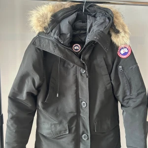 Svart parkajacka från Canada Goose - Säljer en svart parkajacka från Canada Goose med pälskantad huva och klassisk logga på ärmen. Jackan har dragkedja, knappar och flera fickor framtill samt en ficka på ärmen. Jackan är XS men passar S