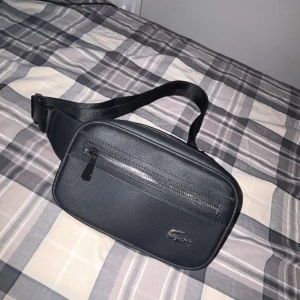 Svart magväska från Lacoste - Snygg svart magväska från Lacoste med justerbart band och två dragkedjefack. Väskan har en stilren design med Lacoste-loggan i metall på framsidan. Perfekt för att bära det viktigaste och hålla händerna fria. Materialet är tåligt och lätt att torka av.