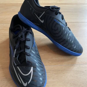 Nike Phantom svart/blå fotbollsskor - Säljer ett par Nike Phantom gx fotbollsskor för inomhusspel. Svarta med blå sula och silverfärgade detaljer. Skorna har snörning och ett mönster över hela ovandelen. Perfekta för inomhusspel med sin platta, greppvänliga sula och sportiga look.
