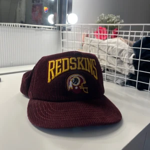 Vinröd Redskins keps i manchester - Säljer en vinröd snapback-keps från New Era med Redskins-logga broderad i gult framtill. Kepsen är tillverkad i manchestertyg och har justerbar baksida. Klassisk retrostil med markerad skärm och cool sportig vibe.