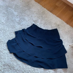 Mörkblå volangkjol från Vero Moda - Supersöt mörkblå kjol från Vero Moda med bred smock i midjan och flera lager volanger. Kjolen har en kort och flowy passform som ger en snygg siluett. Perfekt att styla med en enkel topp eller stickad tröja.