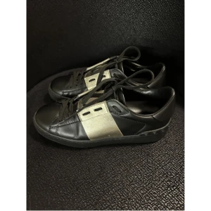 Valentino Garavani sneakers svart/guld - Snygga svarta sneakers från Valentino Garavani med bred guldfärgad panel på sidorna och coola nitar baktill på sulan. Köpta på Vestiaire Collective 