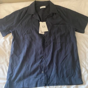 Mörkblå kortärmad skjorta These Glory Days - Snygg mörkblå skjorta från These Glory Days i storlek S. Skjortan har korta ärmar, klassisk krage, bröstficka och knäppning framtill. Tillverkad i mjukt och svalt tencel-material som ger en skön känsla. Perfekt för dig som gillar stilren och enkel design.