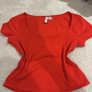Röd topp med rund hals från H&M - En enkel och snygg röd topp från Divided by H&M. Modellen har korta ärmar och en djup rundad halsringning. Tillverkad i mjuk bomull som känns skön mot huden. Perfekt att matcha med jeans eller kjol för en chill look.