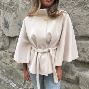 Beige cape  - Populär beige cape med knytbälte i midjan och vida trekvartsärmar. Hög krage med dekorativ guldfärgad knapp framtill. Perfekt för dig som vill ha en elegant och trendig look.