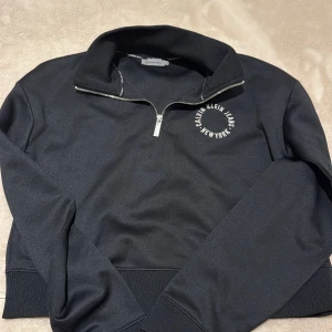 Svart Calvin Klein half zip sweatshirt - Svart sweatshirt från Calvin Klein Jeans med halv dragkedja och broderad logga på bröstet. Tröjan har hög krage, ribbade muddar och är i ett mjukt material som känns skönt mot huden. Perfekt för en chill och stilren look.