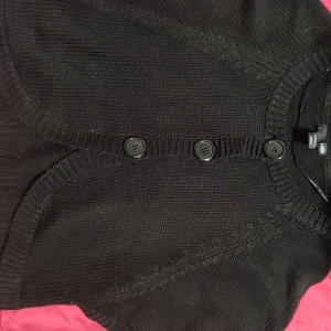 Svart kofta från H&M med knappar - Svart stickad kofta från H&M med tre stora knappar framtill. Koftan har ribbade detaljer vid halsringning och knappslå, samt korta ärmar. Perfekt att ha det över en långärmad tröja.