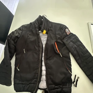  Parajumpers - Svart pufferjacka från Parajumpers med coola detaljer som orange dragkedjor, gul PJS-rem vid kragen och patch på ärmen. Jackan har flera fickor med dragkedja och quiltad design. Perfekt för dig som vill ha en snygg och funktionell jacka med streetkänsla. dragkedjan sönder