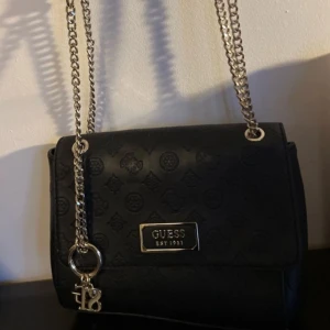 Svart axelväska från Guess med kedja - Snygg svart axelväska från Guess med präglat logomönster och silverfärgad kedjerem. Väskan har metallplatta framtill med Guess-logga och dekorativa berlocker. Tillverkad i skinnimitation och har en stilren, fyrkantig form.