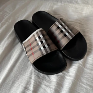 Burberry tofflor - Snygga sandaler med klassiskt Burberry-rutigt mönster i beige, svart, vitt och rött. De har en bred rem över foten och svart platt sula i syntetmaterial. Perfekta för sommaren och riktigt trendiga att glida runt i.