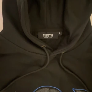 Trapstar London Shooters svart hoodie - Svart hoodie från Trapstar med stor broderad London Shooters-logga i grått, blått och vitt framtill. Klassisk känguruficka, snörning i huvan och ribbade muddar. Perfekt streetwear-vibe och riktigt snygg detalj med pantermotiv.