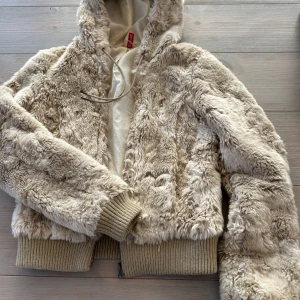 Beige pälsjacka med huva från H&M Divided - Mysig beige pälsjacka från H&M Divided i storlek 36. Jackan har huva, ribbade muddar och dragkedja framtill. Perfekt för dig som vill ha en fluffig och trendig look under kyliga dagar.