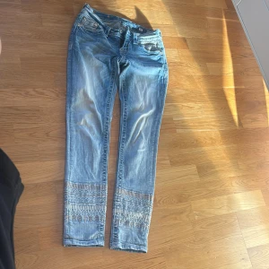 Blå broderade jeans från Miss Me - Snygga ljusblå jeans från Miss Me med broderade detaljer och pärlor längst ner på benen. Jeansen har raka ben, dekorativa sömmar och coola knappar med blommotiv. Perfekt för dig som vill sticka ut med unika detaljer.