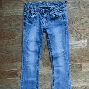 Ljusblåa lowrised bootcut jeans y2k med broderi på fickorna  - Ljusblå jeans från Wallflower med bootcut passform och slitna detaljer framtill. Snygga broderade bakfickor med knappar och klassisk femficksdesign. Jeansen är tillverkade i denim och har en cool, avslappnad vibe. Storlek Xs yttersömn 94cm, innersömn 70cm, midjebredd 34cm)