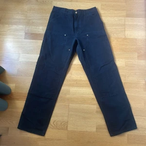 Carhartt Double Knee Pant svart - Svart Double Knee Pant från Carhartt med raka ben, förstärkta knän och flera praktiska fickor. Byxorna har klassisk Carhartt-logga på bakfickan och är tillverkade i slitstark bomull. Perfekta för dig som gillar workwear-stil och funktionella detaljer.