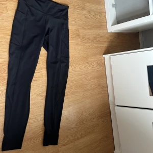 Svarta leggings med fickor - Svarta leggings från CRIVIT med hög midja och praktiska sidofickor. Tillverkade i stretchigt syntetmaterial som sitter tajt och bekvämt. Perfekta för träning eller chill dagar. Snygg och enkel design som passar till det mesta.