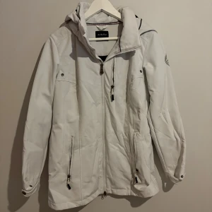 Vit vindjacka från On The Peak - Snygg vit vindjacka från On The Peak med huva, dragkedjor framtill och på fickorna samt coolt broderat märke på ärmen. Jackan har en sportig look och är perfekt för blåsiga dagar. Passformen är normal och jackan har långa ärmar.