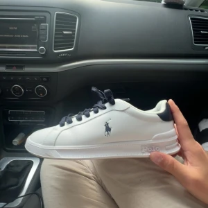 Vita Polo Ralph Lauren sneakers - Snygga vita sneakers från Polo Ralph Lauren med mörkblå detaljer och snörning. Klassisk låg modell med Polo-logga på sidan och bekväm platt sula. Perfekta för en clean och stilren look.