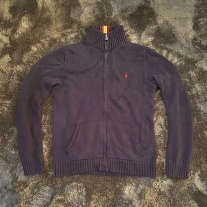 Mörkblå stickad kofta från Polo Ralph Lauren - Mörkblå stickad kofta från Polo Ralph Lauren med dragkedja och ribbad krage med röda och gula ränder. Klassisk röd logga broderad på bröstet och två fickor framtill. Perfekt för lager-på-lager under kyliga dagar. Size 18-20 år passar S