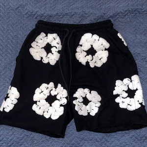Svarta shorts från Denim Tears med blommor - Svarta shorts från Denim Tears med vita blommiga tryck över hela plagget. De har elastisk midja med snörning 
