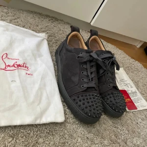 Christian Louboutin svarta sneakers med nitar - Svarta sneakers från Christian Louboutin i mocka med ikonisk röd sula och svarta nitar på tån. Skorna har snörning och rund tå, samt detaljer i skinn på insidan. Snygg och edgy design som sticker ut. Dustbag och kvitto följer med men ingen låda. En nit är borta också
