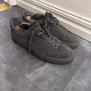 Mörkgrå sneakers från Garment Project strl 44 - Snygga mörkgrå sneakers från Garment Project i mocka med klassisk låg profil och rund tå. Skorna har matchande mörkgrå snörning och platt sula, vilket ger en clean och stilren look. Perfekta för dig som gillar minimalistisk design och vill ha något som funkar till det mesta.