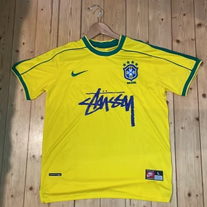 Brasilien x Stüssy x Nike fotbollströja L - Storlek L men passar även M. Gul Brasilien fotbollströja i samarbete med Stüssy och Nike. Tröjan har gröna detaljer, broderat CBF-märke, Nike-logga och Stüssy-tryck på bröstet. Tillverkad i lätt och ventilerande polyester med Dri-FIT-teknologi. Perfekt för dig som gillar streetwear och fotboll.
