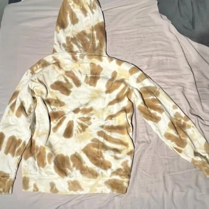 Vans tie dye hoodie med tryck - Säljer en beige och vit tie dye hoodie från Vans med coolt tryck på bröstet och snörning i huvan. Mjuk bomullskänsla och avslappnad passform, perfekt för chill dagar. Unik färgkombination som sticker ut.