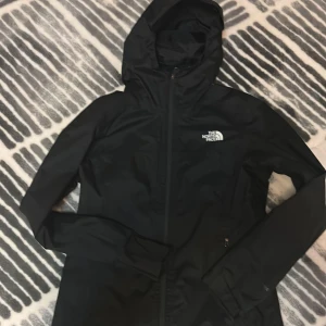 Svart skaljacka / regnjacka från The North Face - Svart regnjacka / skaljacka från The North Face med huva och dragkedja framtill. Jackan har meshfoder på insidan, fyra fickor med dragkedja och logga på bröstet. Perfekt för blåsiga och regniga dagar och enkel att matcha med allt. Jackan har inga skador och är knappast använd. Pris kan diskuteras.
