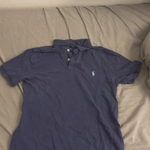 Marinblå Polo Ralph Lauren t-shirt - Säljer en marinblå t-shirt från Polo Ralph Lauren i slim fit-modell. Klassisk piké med korta ärmar, krage och knappar framtill. Ikoniska loggan broderad på bröstet. Tillverkad i mjuk bomull som känns skön mot huden. Det står M men skulle säga att det är en S
