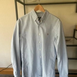 Ralph Lauren skjorta - Klassisk ljusblå skjorta från Polo Ralph Lauren i slim fit i storlek M. Skjortan har button down-krage, broderad logga på bröstet och knäppning framtill. Tillverkad i mjuk bomull och har långa ärmar med manschettknappar. Perfekt för en clean och stilren look.