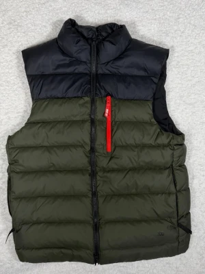 Nike SB dunväst i svart och olivgrön - Snygg dunväst från Nike SB i svart och olivgrön med en röd dragkedja på bröstfickan. Västen har quiltad design, hög krage och hel dragkedja framtill. Perfekt för lager-på-lager och streetwear. Märke och storlek syns tydligt på insidan.