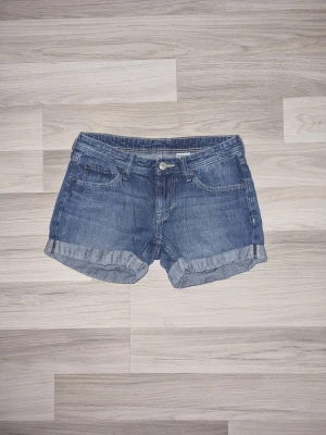 Blå jeansshorts från H&M - Säljer ett par klassiska blå jeansshorts från H&M med upprullade benslut och femficksmodell. Shortsen har normal passform och är tillverkade i slitstark bomull. Perfekta för sommardagar och enkla att matcha med allt.