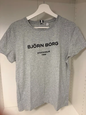 Grå t-shirt från Björn Borg - Snygg ljusgrå t-shirt från Björn Borg med svart tryck på bröstet där det står 'BJÖRN BORG STOCKHOLM 1956'. Tillverkad i 100% BCI bomull och har klassisk rund hals och korta ärmar. Perfekt till en avslappnad stil.