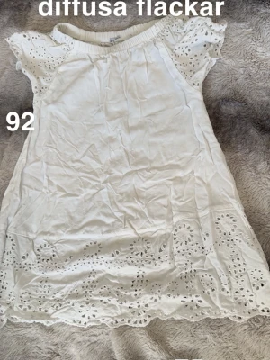 Vit kortklänning med spetsdetaljer - Söt vit kortklänning med broderade spetsdetaljer på ärmar och nederkant. Klänningen har korta ärmar och en avslappnad passform, perfekt för varma dagar. 🌸Diffusa fläckar. Storlek 92