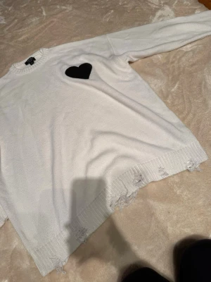 Vit stickad tröja med svart hjärta BoohooMAN - Säljer en vit stickad tröja från BoohooMAN i storlek M. Tröjan har ett svart hjärta på bröstet och slitna detaljer längst ner. Långärmad och med ribbade muddar vid hals, ärmslut och nederkant. Perfekt för dig som gillar streetstyle och unika detaljer.