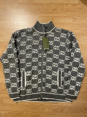 Grå stickad kofta med GG mönster  - Säljer en grå stickad kofta  med hel dragkedja och hög krage. Tröjan har ett vitt GG-mönster över hela plagget, ribbade muddar och fickor framtill. har snygga vita detaljer vid krage, ärmslut och nederkant. Passar storlek L