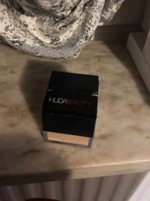 Huda Beauty lös puder i färgen banana bread  - Säljer ett löspuder från Huda Beauty i en fyrkantig burk med svart lock och transparent botten. Pudret har en ljus beige ton som ger en matt finish och passar perfekt för att fixera din bas. Snygg och stilren förpackning som är enkel att använda.