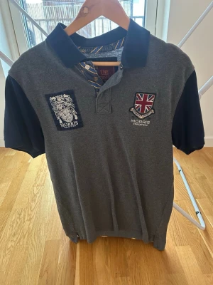 Grå piké från Morris Tradition - Säljer en grå pikétröja från Morris Tradition med mörkblå krage och ärmar. Tröjan har broderade patchar på bröstet, en med brittisk flagga och en med Morris-logga. Klassisk pikéknäppning och snygga detaljer vid kragen.