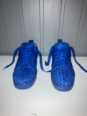 Blå mocka sneakers med nitar - Säljer ett par riktigt snygga blå sneakers från Christian Louboutin i mocka med matchande blå snörning och coola nitar över tån. Skorna har den klassiska röda sulan och en låg profil som ger en clean look. Perfekta för dig som vill sticka ut med stil. Ingen box tyvärr.