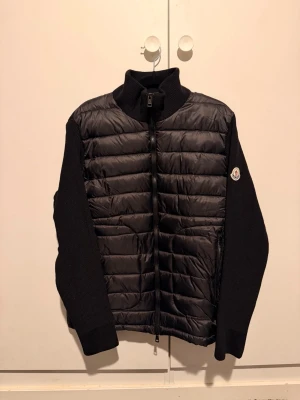 Moncler cardigan  - Säljer en svart Moncler Cardigan med stickade ärmar och quiltad front. Jackan har hög krage, dragkedja framtill och Moncler-logga på ärmen. Perfekt för dig som vill ha en clean och stilren look med premiumkänsla.