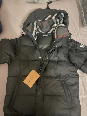 Svart pufferjacka från Burberry - Säljer en svart pufferjacka från Burberry med huva och klassiskt rutigt foder i grått, vitt och rött. Jackan har dragkedja, knappar och två fickor med dragkedja. På ärmen sitter en Burberry London England-patch. Perfekt för kalla dagar.