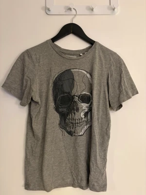 Dam Grå t-shirt med dödskalletryck - Snygg grå t-shirt med ett stort dödskalletryck på framsidan från en affär utomlands i storlek 164cm.  Klassisk rund halsringning och korta ärmar. Perfekt för dig som gillar edgy och avslappnad stil.