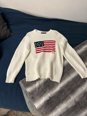 Vit stickad tröja Polo Ralph Lauren - Säljer en vit stickad tröja från Polo Ralph Lauren med amerikansk flagga i rött, blått och vitt på bröstet. Tröjan har rund hals, ribbade muddar och RL-broderi. Perfekt för dig som gillar klassisk och fancy stil.